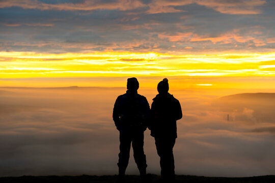 Anonymous couple standing on mountain together in love watching sunrise silhouetted stood high above the clouds looking down valley with golden hour dawn sunlight on the horizon wrapped up warm