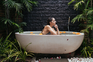 Spa surrounded by palm trees. at the stone wall stands a bath with water and buds of flowers in it sits a girl in a swimsuit propped up her chin. High quality photo - 416095817