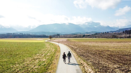 A panoramic, drone shot of a couple walking on a road leading to the mountains an alpine landscape of Austria. Lush green meadows and crop fields spread on a vast surface. Idyllic landscape.
