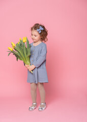 beautiful little blonde girl in blu dress with bouquet of yellow tulips on pink background with...