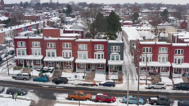 Aerial of colorful row houses covered in snow, decorated for holidays. American inner city during winter. Cars parked along snowy street.