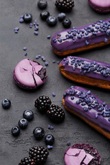 Set of purple eclairs, macarons, blueberries and blackberries on dark concrete background. Side view, close up
