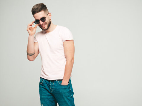 Portrait of handsome smiling stylish hipster lambersexual model.Man dressed in white T-shirt and jeans. Fashion male posing on grey background in studio in sunglasses