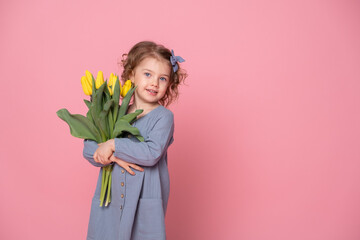beautiful little blonde girl in blu dress with bouquet of yellow tulips on pink background with...