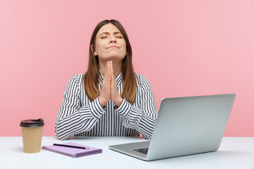 Hopeful brunette woman pressing hands and praying heartily, keeping eyes closed, asking god for help, sitting at workplace. Indoor studio shot isolated on pink background
