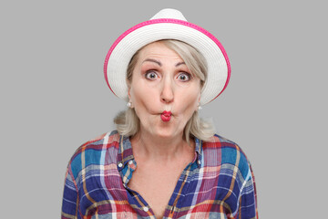Portrait of funny shocked modern stylish mature woman in casual style with white hat standing with fish lips and looking at camera with crazy face. indoor studio shot isolated on gray background.