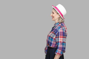 Profile side view portrait of happy successful modern stylish mature woman in casual style with hat standing, looking forward with toothy smile. indoor studio shot isolated on gray background.