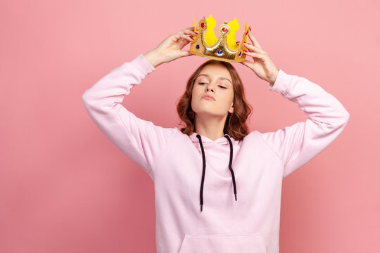 Portrait of selfish curly haired teenage girl in hoodie holding gold crown over head, looking with arrogance, privileged status. Indoor studio shot isolated on pink background