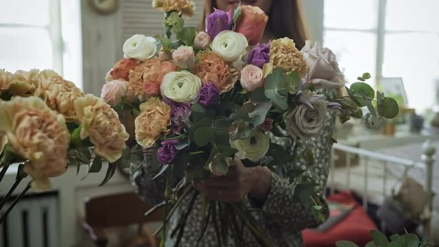 Female self employed florist is creating beautiful flower arrangement in vintage workshop. Young woman is putting bunch of fresh roses carnations and peonies in bouquet