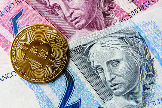 Electronic golden bitcoin coin with a brazilian real at the background. Devaluation of the Brazilian real against the decentralized currency.