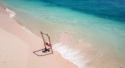 Fototapeta na wymiar A girl swinging on a swing placed on the seashore of Pink Beach, Lombok, Indonesia. The swing has simple wood construction. Waves wash the pillars of it. In the back there are few boats. Drone capture