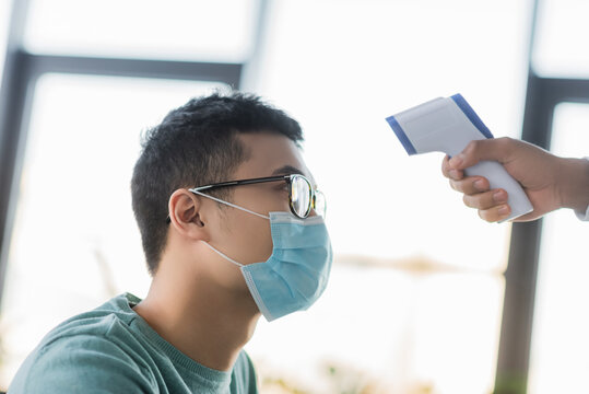 Asian businessman in medical mask near colleague with non-contact thermometer