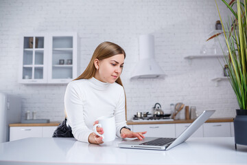 Portrait of lovely young woman studying at home on laptop. A girl with long blond hair in a sweater drinks tea. Modern kitchen interior and education concept. Work remotely from home