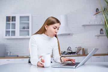 A beautiful young woman with blond hair in a white turtleneck sweater stands in a white kitchen and works at a laptop, she drinks coffee. Work remotely from home