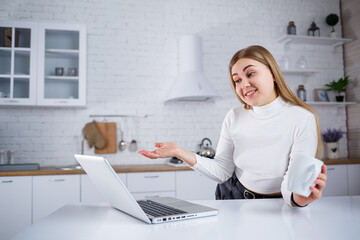 Obraz na płótnie Canvas Beautiful young woman studying at home on a laptop. A girl with long blond hair in a sweater drinks tea sits in the kitchen. Work remotely from home