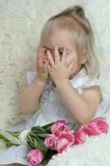 girl of two years old, blonde is sitting in the room on the couch with a bouquet of pink tulips, covered her eyes with her hands, concept small woman, birthday, happy childhood