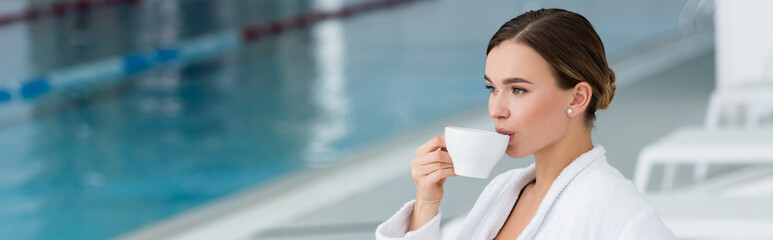 Young woman in bathrobe drinking tea near swimming pool on blurred background, banner