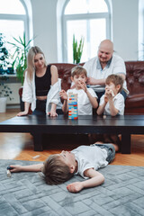 Cosy and modern living room. Cheerful boy lying on floor while his brother and parents play jenga game together.