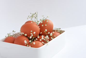 Easter eggs in a white square dish, on a white table, with a sprig of delicate flowers. Close-up, space for text