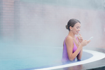 happy woman in swimsuit holding smartphone while bathing in outdoor swimming pool in spa center