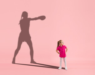Childhood and dream about big and famous future. Conceptual image with girl and drawned shadow of professional female boxer on coral pink background. Childhood, dreams, imagination, education concept.