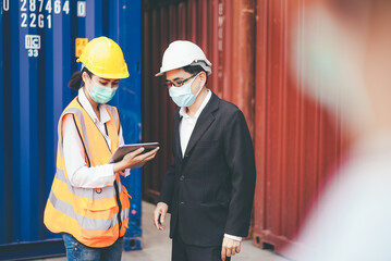 Asian logistic foreman wearing protective mask working with female worker showing report from digital tablet. import export shipping business. freight cargo and container warehouse concept