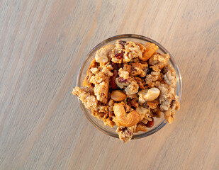 Overhead view of a small glass bowl filled with whole grains granola with fruit and nuts on a table illuminated with natural light.