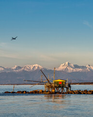 An airplane flies over a trebuchet fishing hut at sunset, against the snow-capped Alps, Marina di...