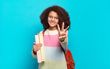 pretty afro teenager smiling and looking friendly, showing number three or third with hand forward,...