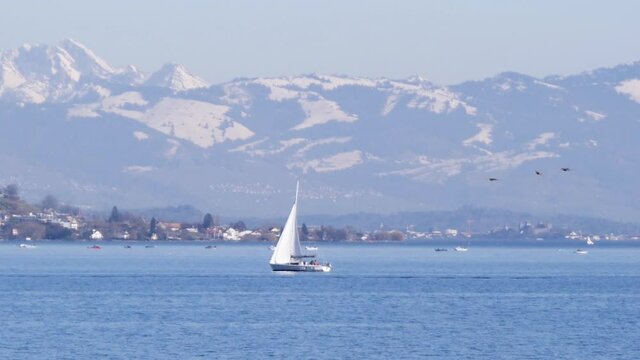 Slow motion sailboat on European lake with big snowy mountains.