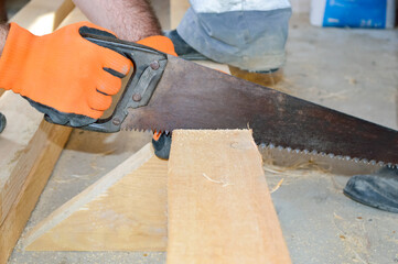 close-up - a man sawing a wooden board with a saw, his hands in orange protective gloves; the assistant holds the board with his feet