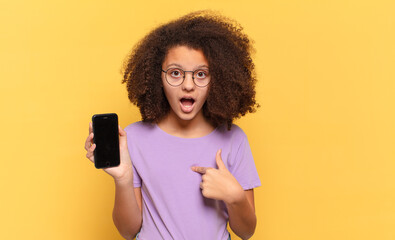 pretty afro teenager looking shocked and surprised with mouth wide open, pointing to self and...