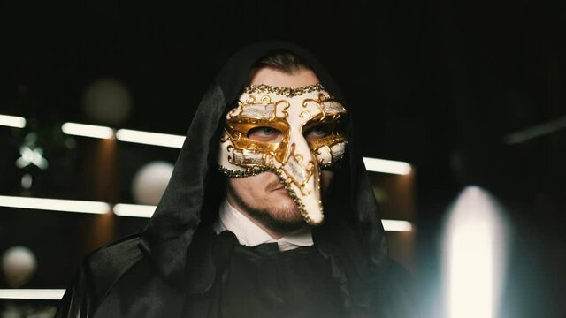 lonely mystical man actor in a black robe and with a hood on his head wearing a Venetian mask stands in a luxurious place with backlighting in the background and looks around