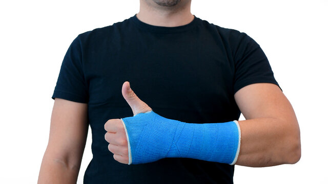 Unrecognizable man in a black T-shirt shows thumbs up and hand wrapped in a fiberglass plaster cast on a white background. Broken wrist in modern blue waterproof bandage