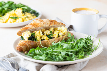 Delicious croissant sandwich with scrambled spinach eggs served with arugula on plate and coffee...