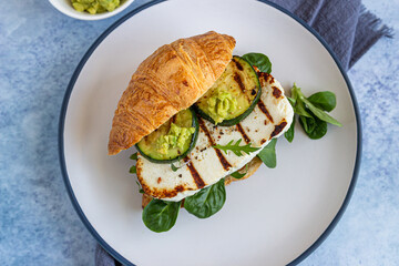 Croissant sandwich with grilled halloumi cheese, arugula and grilled zucchini, blue concrete background. Healthy breakfast or lunch.