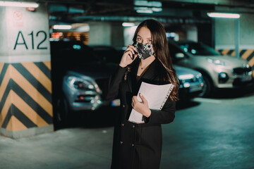 Young happy businesswoman with pandemic mask communicating over mobile phone in underground parking.