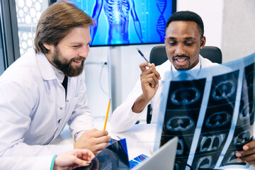 Fototapeta premium Young male doctors discuss the results of an MRI scan of a patient in the hospital, wearing white coats with stethoscopes. Medicine and healthcare concept.