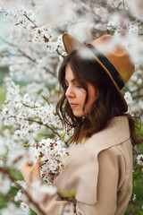 a woman in a beige coat and hat stands in a park or garden near a blossoming apple tree