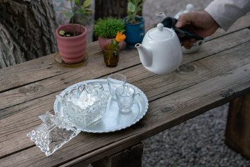 Pouring traditional Iranian tea. White teapot, crystal sugar, plate on a wooden table outdoors. Shiraz, Iran.