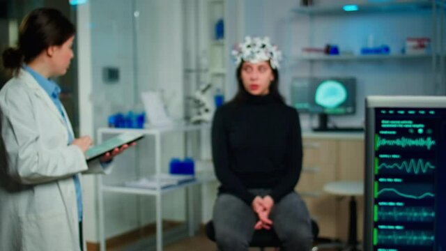 Doctor specialist in neurological medicine typing health informations of patient. Woman wearing eeg headset answering to medical researcher, monitor showing brain scan in equipped laboratory