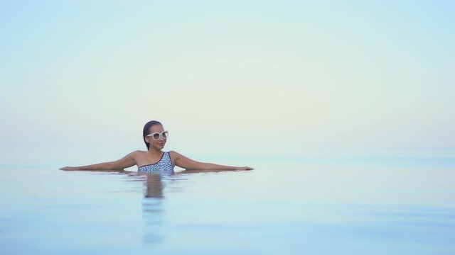 A woman with her arms spread out along the edge of an infinity pool appears to be floating in space against an ocean horizon. Title space