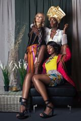 Portrait of a group of 3 elegant women with curly long hair and beautiful makeup posing by themselves in a studio with curtain background wearing designer clothing and high heels.