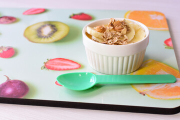 Chopped banana with granola in white bowl, on wooden background printed with fruits