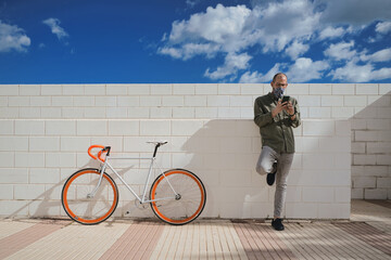 Adult man wearing face mask using his mobile phone next to orange bike against white wall.