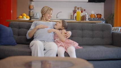 Happy, peaceful family concept.Loving little girl loving her belly with her hands chatting with her pregnant mother while sitting on the sofa. Slow motion video.