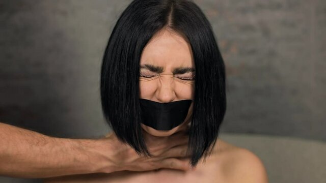 Sad brunette woman with duct tapered mouth grabbed by male hands over neck in domestic violence concept