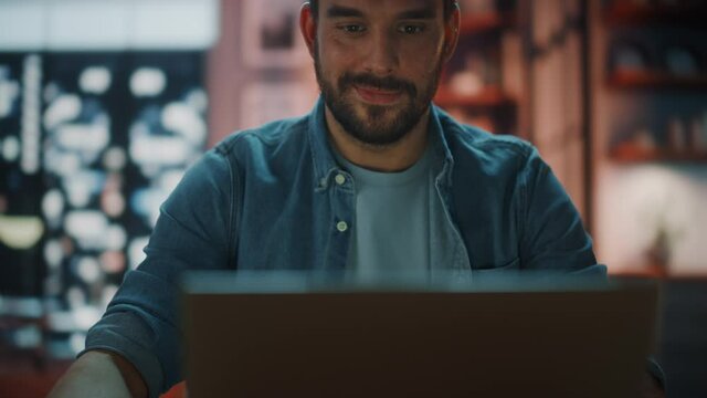 Handsome Caucasian Man Working on Laptop Computer while Sitting at a Desk in Dark Cozy Living Room in the Evening. Freelancer Working From Home. Browsing Internet, Using Social Networks, Having Fun.
