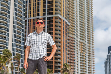 Portrait of young handsome attractive man hipster millennial on beach with red sunglasses in Miami, Florida with hands on hips by apartment condo building