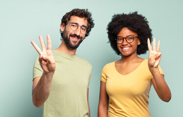 multiracial couple of friends smiling and looking friendly, showing number three or third with hand...